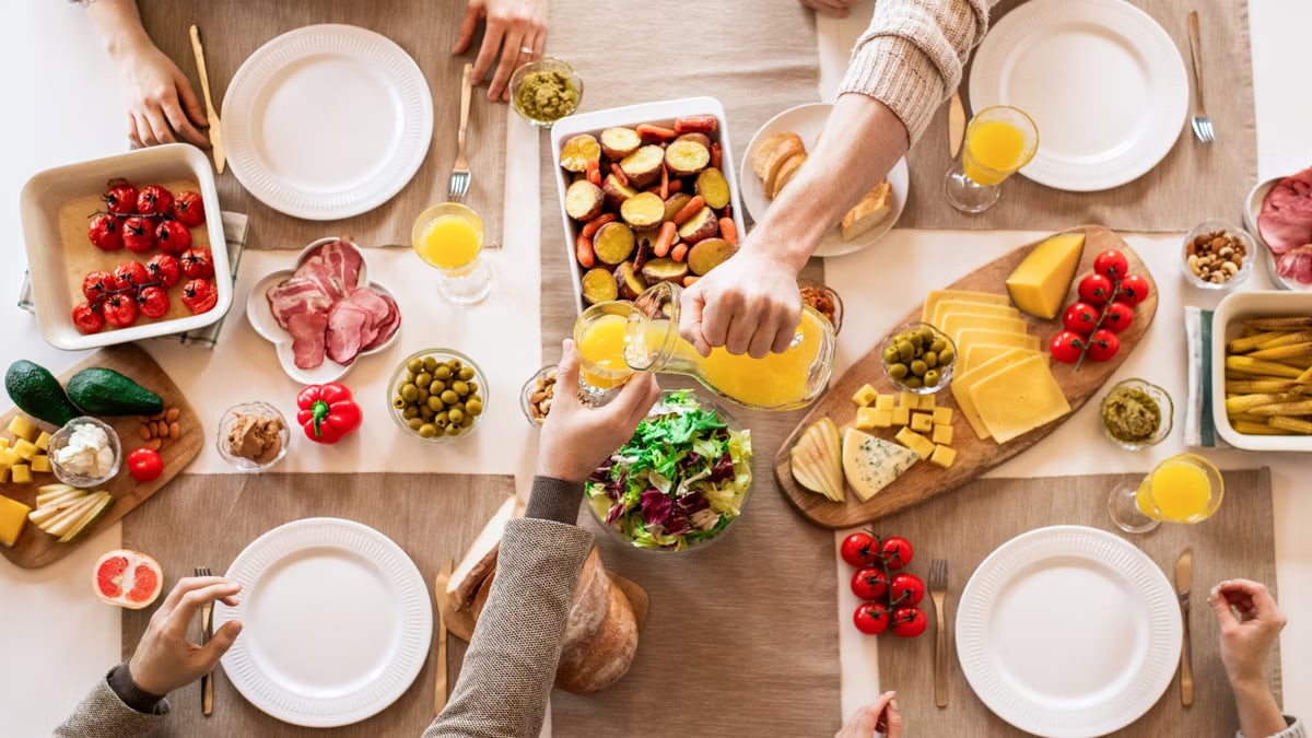 Overhead view of a lively breakfast table featuring orange juice, cheeses, fresh vegetables, and cured meats.
